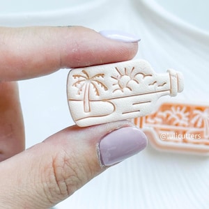 May include: A white plastic cookie cutter shaped like a bottle with a palm tree, sun, and clouds inside. The cutter is being held by a hand.