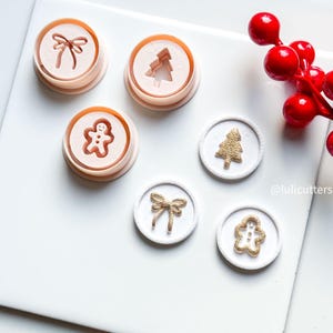 以下が含まれることがあります： クリスマスをテーマにしたクッキーカッターのコレクション。リボン、クリスマスツリー、ジンジャーブレッドマンのデザインが含まれています。カッターはピーチ色で、白い表面に置かれています。金色のテクスチャードクッキーの装飾も表示されています。