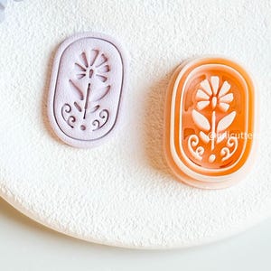 May include: Two floral-shaped cookie cutters. One is lavender, the other is orange. Both are oval-shaped with a flower design in the centre. The orange cutter has the text "@licutters" on the side. The cutters are on a white surface.