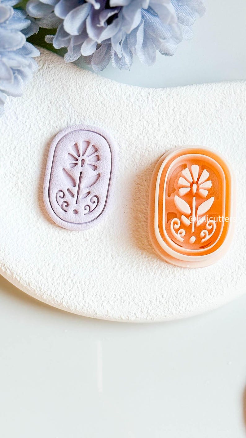 May include: Two floral-shaped cookie cutters. One is lavender, the other is orange. Both are oval-shaped with a flower design in the centre. The orange cutter has the text "@licutters" on the side. The cutters are on a white surface.