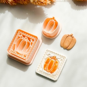 May include: A collection of pumpkin-themed cookie cutters and a decorated cookie. The cutters are in various shapes and sizes, including a layered square cutter and a single pumpkin shape. The decorated cookie features an orange pumpkin design on a white background.