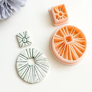 Könnte beinhalten: Eine Sammlung von Bastelwerkzeugen, darunter zwei quadratische und ein kreisförmiger Ausstecher in Hellorange. Außerdem gibt es zwei Keramikteile mit einem radialen Design in Weiß und Grün. Die Artikel sind auf einer weißen Oberfläche angeordnet.