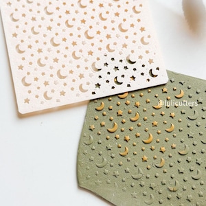 May include: Two stencils with celestial designs. One is a light beige square with a pattern of crescent moons and stars. The other is an olive green, irregularly shaped stencil with the same moon and star pattern. The stencils are for crafting.