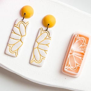 Pode incluir: Um par de brincos pendentes brancos e amarelos com um design floral. Os brincos são feitos de argila polimérica e têm um gancho de metal dourado. Também é mostrado na imagem um molde de silicone rosa com o mesmo design floral.