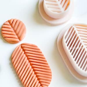 May include: Two silicone moulds for making polymer clay leaf shapes. The moulds are pink and feature a leaf design with raised lines. There are two orange polymer clay leaves, one round and one oval, that have been pressed into the moulds.