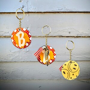 May include: Three keychains with different designs. The first keychain has a red and orange circle with a white letter "B" in the center. The second keychain has a red and orange circle with a yellow letter "I" in the center. The third keychain has a yellow circle with a black smiley face in the center.