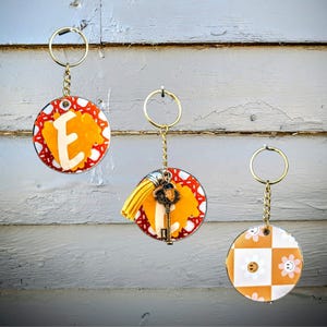 May include: Three keychains with different designs. The first keychain has a round orange and red patterned charm with the letter "E" in the center. The second keychain has a round orange and red patterned charm with a small key charm. The third keychain has a round orange and white checkered charm with a smiley face.
