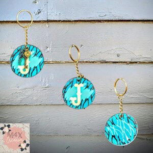 May include: Three turquoise blue keychains with a gold key charm and a white letter on each. The keychains have a black and white animal print design.