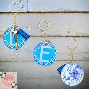 May include: Three round keychains with gold-colored hardware. Two keychains have a light blue background with a white letter and a decorative border. One keychain has a blue tassel and a gold-colored key charm. The third keychain has a blue and white marbled design.