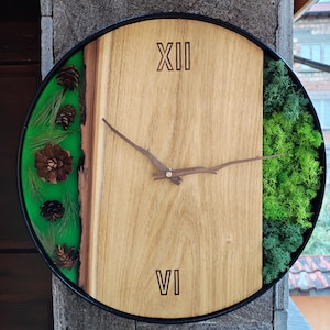 May include: A round wooden clock with Roman numerals, a black frame, and a green epoxy resin section with pine cones. The clock face is made of wood with a natural wood grain.