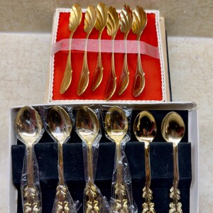 May include: A set of gold-colored spoons, some with a leaf design on the handle, and others with a swirl design on the spoon head. The spoons are arranged in two separate boxes, one with a red background and the other with a black background.