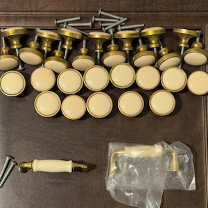 May include: A collection of vintage cabinet hardware, including brass and cream-colored knobs and pulls. The knobs have a classic design with a brass base and a round, cream-colored top. Also included are screws for installation.
