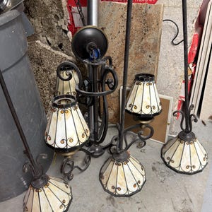 May include: A collection of antique-style chandeliers and lamps. The lamps feature off-white shades with a stained glass design, accented with dark metal frames. The fixtures are ornate, with curved arms and decorative elements.