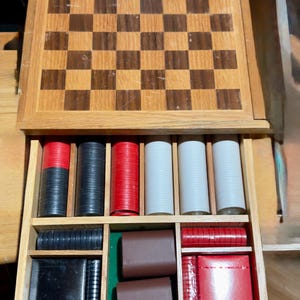 May include: Wooden chess and checkers set with a drawer containing game pieces. The board is brown and tan with a checkerboard pattern. The drawer holds red, black, and white game pieces, along with brown leather-like pieces.