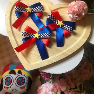 Pu&ograve; includere: Tre fiocchi per capelli rossi, blu e a scacchi con accenti a stella gialla sono disposti su un vassoio di legno a forma di cuore. Nel'immagine sono visibili anche un fiore rosa e una bambola di peluche.