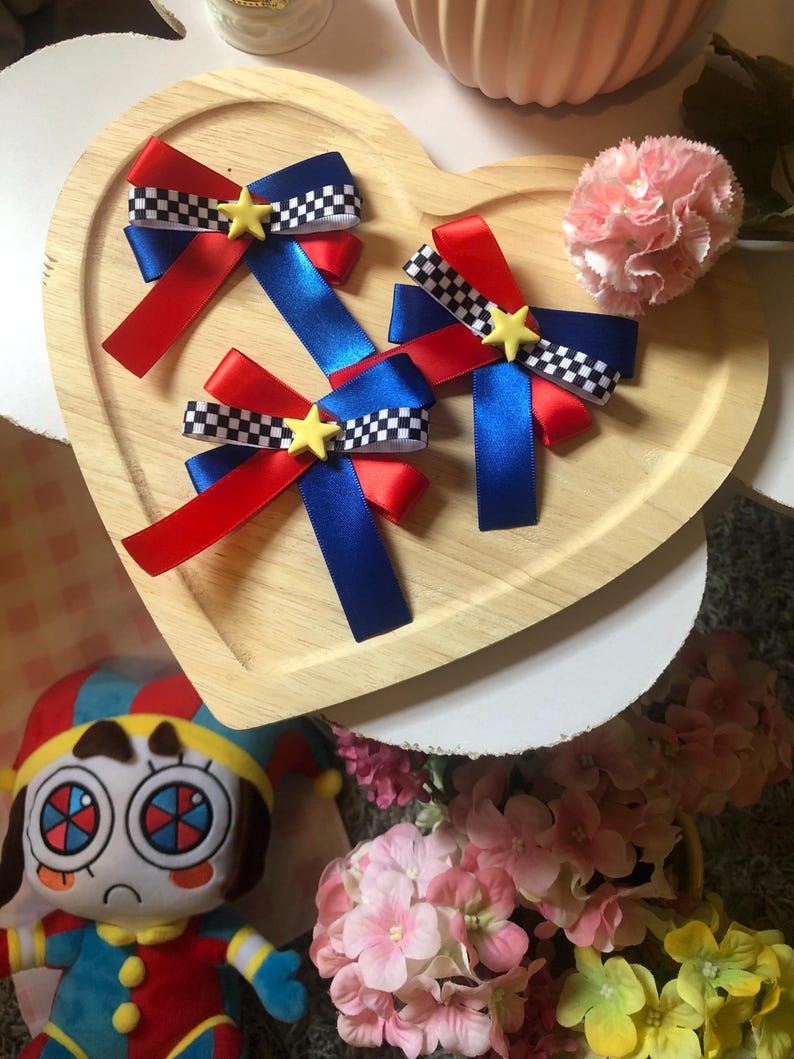 Pu&ograve; includere: Tre fiocchi per capelli rossi, blu e a scacchi con accenti a stella gialla sono disposti su un vassoio di legno a forma di cuore. Nel'immagine sono visibili anche un fiore rosa e una bambola di peluche.
