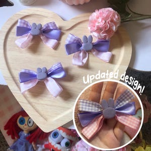 Pu&ograve; includere: Tre fiocchi per capelli fatti a mano con un centro a forma di coniglietto. I fiocchi presentano nastri viola e rosa, con un motivo a quadretti su alcuni. I fiocchi sono esposti su un vassoio di legno a forma di cuore. Il testo "Updated design!" &egrave; visibile.