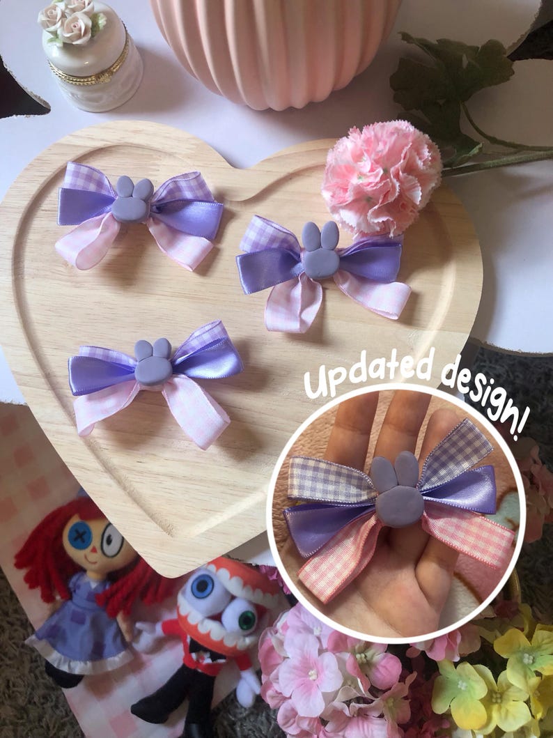 Pu&ograve; includere: Tre fiocchi per capelli fatti a mano con un centro a forma di coniglietto. I fiocchi presentano nastri viola e rosa, con un motivo a quadretti su alcuni. I fiocchi sono esposti su un vassoio di legno a forma di cuore. Il testo "Updated design!" &egrave; visibile.