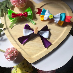 Pu&ograve; includere: Tre fiocchi colorati per capelli su un vassoio di legno a forma di cuore. I fiocchi presentano vari colori tra cui verde, rosso, viola, bianco, giallo e blu. Un fiocco ha un piccolo accento floreale giallo.