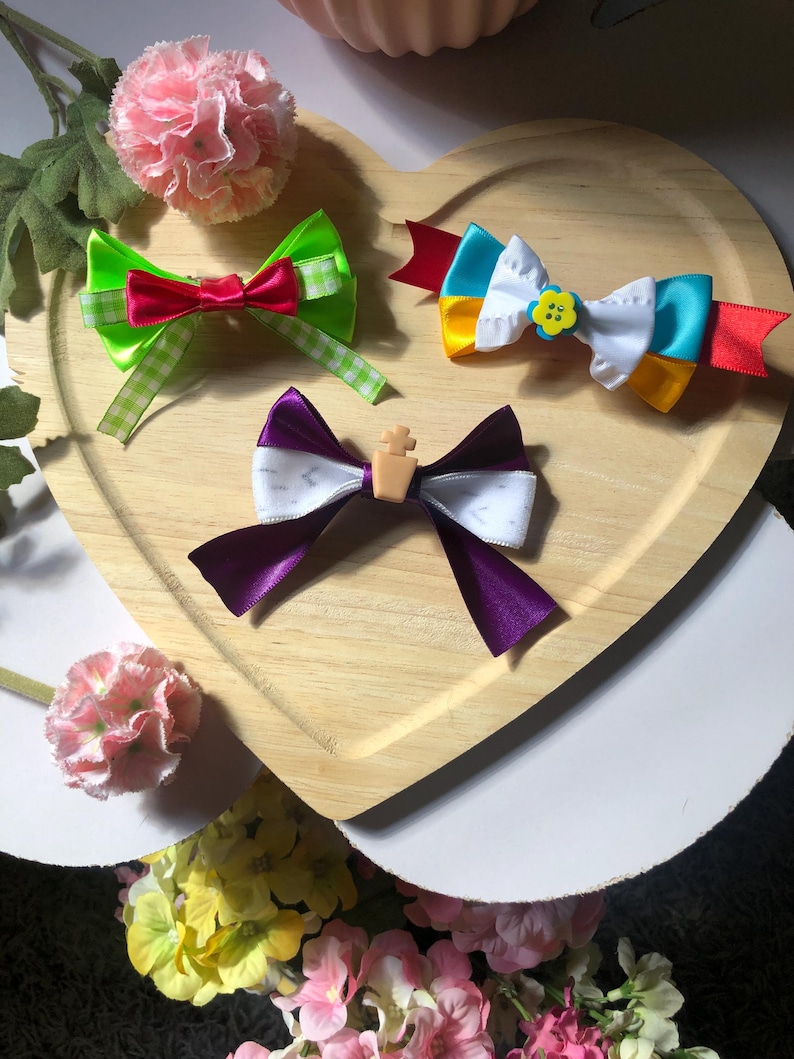 Pu&ograve; includere: Tre fiocchi colorati per capelli su un vassoio di legno a forma di cuore. I fiocchi presentano vari colori tra cui verde, rosso, viola, bianco, giallo e blu. Un fiocco ha un piccolo accento floreale giallo.