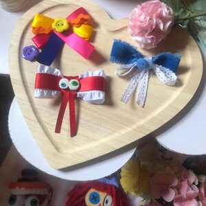 Pu&ograve; includere: Un vassoio di legno a forma di cuore mostra fiocchi per capelli colorati e un garofano rosa. I fiocchi presentano vari colori come giallo, rosa, rosso e blu, con bottoni e nastri. Due bambole sono visibili in basso.