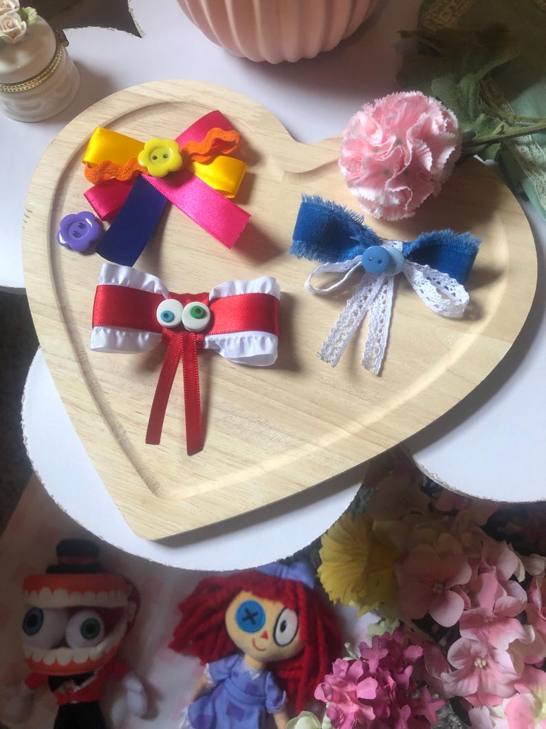 Pu&ograve; includere: Un vassoio di legno a forma di cuore mostra fiocchi per capelli colorati e un garofano rosa. I fiocchi presentano vari colori come giallo, rosa, rosso e blu, con bottoni e nastri. Due bambole sono visibili in basso.
