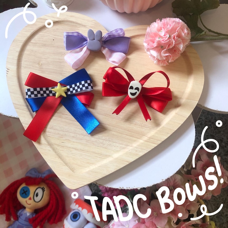 Pu&ograve; includere: Tre fiocchi decorativi in nastro colorato sono disposti su un vassoio di legno a forma di cuore. Un fiocco &egrave; rosso, blu e bianco con una stella, un altro rosso con un teschio e il terzo viola e rosa con un coniglietto. Il testo "TADC BOWS!" &egrave; visibile.