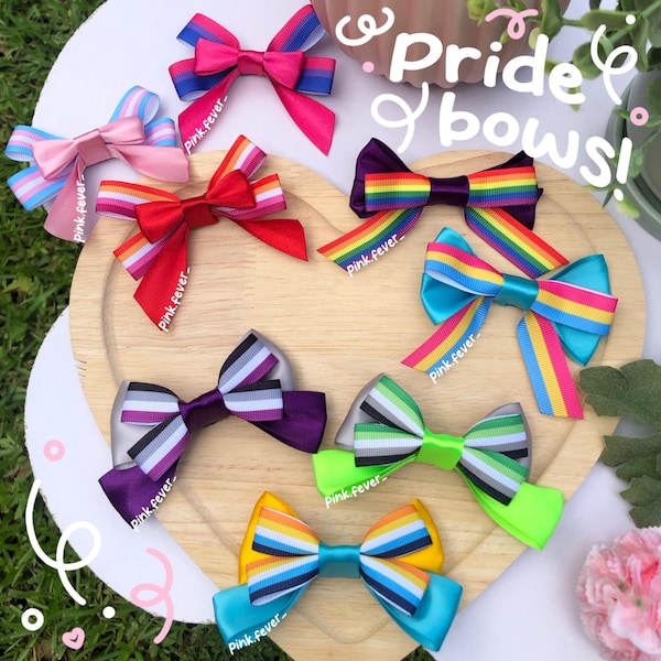 May include: A collection of seven hair bows featuring various pride flag color patterns, including bisexual, lesbian, rainbow, pansexual, asexual, aromantic, and non-binary designs. Each bow is crafted from satin ribbon with distinct color stripes.