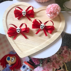 Pu&ograve; includere: Tre fiocchi di raso rosso con centri a forma di teschio bianco su un vassoio di legno a forma di cuore. Un piccolo fiore rosa e altri oggetti decorativi circondano il vassoio. Accessori per capelli.