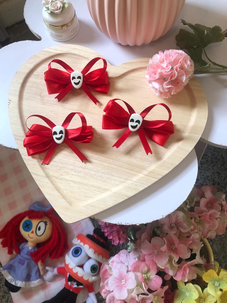 Pu&ograve; includere: Tre fiocchi di raso rosso con centri a forma di teschio bianco su un vassoio di legno a forma di cuore. Un piccolo fiore rosa e altri oggetti decorativi circondano il vassoio. Accessori per capelli.