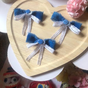 Pu&ograve; includere: Tre fiocchi di jeans blu con dettagli in pizzo bianco e centri con bottoni blu sono disposti su un vassoio di legno a forma di cuore. Un garofano rosa e oggetti decorativi sono sullo sfondo. Accessori per capelli.