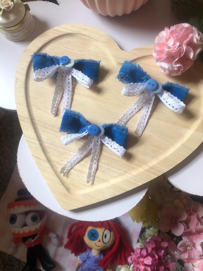 Pu&ograve; includere: Tre fiocchi di jeans blu con dettagli in pizzo bianco e centri con bottoni blu sono disposti su un vassoio di legno a forma di cuore. Un garofano rosa e oggetti decorativi sono sullo sfondo. Accessori per capelli.