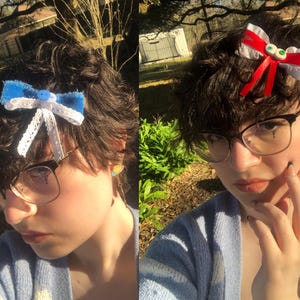 Pu&ograve; includere: Due fiocchi per capelli fatti a mano. Uno &egrave; blu con pizzo bianco e un bottone al centro. L'altro &egrave; rosso e bianco con accenti di occhi verdi. Entrambi sono attaccati a capelli scuri. La persona indossa occhiali e un cardigan azzurro.