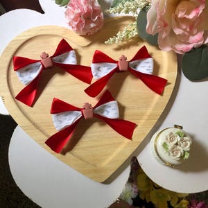 Pu&ograve; includere: Quattro fiocchi di nastro rosso e bianco con un abbellimento a forma di croce sono disposti su un vassoio di legno a forma di cuore. I fiocchi sono circondati da fiori rosa e bianchi e da un piccolo cofanetto decorativo con dettagli di rose.