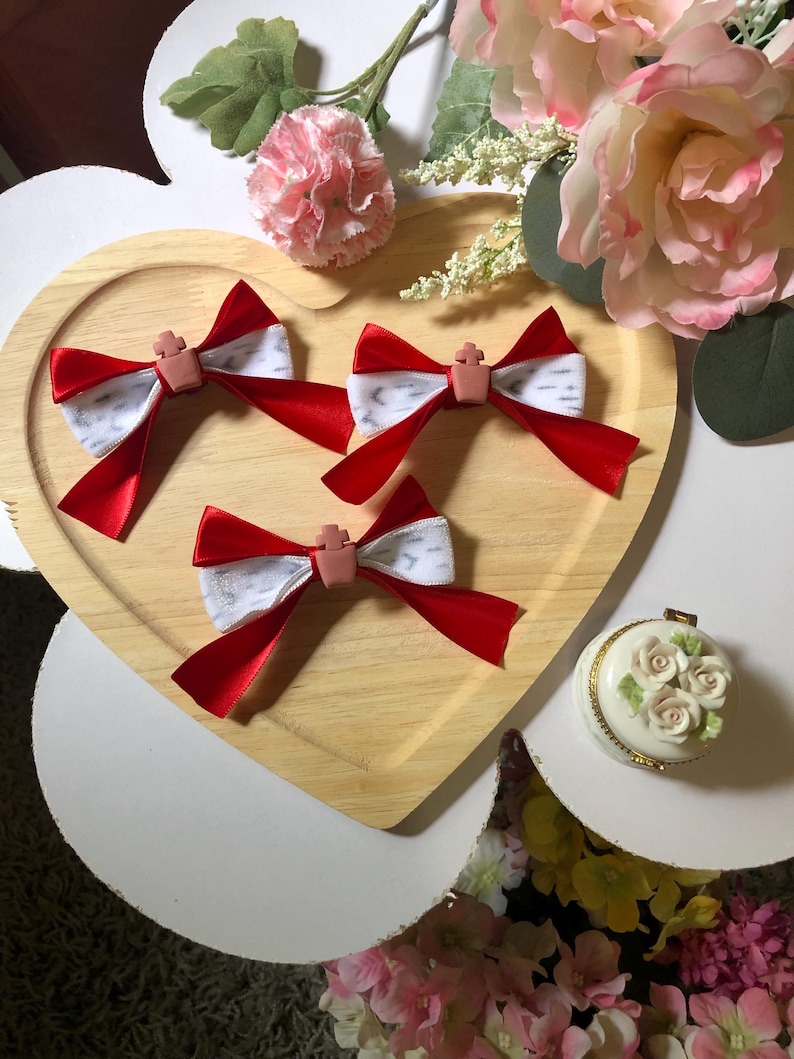 Pu&ograve; includere: Quattro fiocchi di nastro rosso e bianco con un abbellimento a forma di croce sono disposti su un vassoio di legno a forma di cuore. I fiocchi sono circondati da fiori rosa e bianchi e da un piccolo cofanetto decorativo con dettagli di rose.