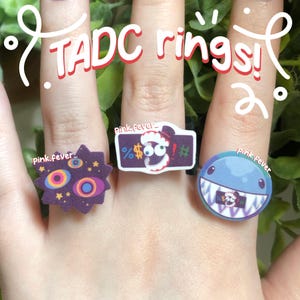 May include: Three rings featuring character designs. The left ring is a purple starburst with colourful eyes. The centre ring is a rectangular purple piece with a character and symbols. The right ring is a round blue face with sharp teeth.