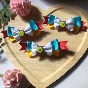 Pu&ograve; includere: Quattro fiocchi per capelli colorati a forma di fiocco, ciascuno con una combinazione di nastri bianchi, gialli e blu turchese, accentuati da una base di nastro rosso e un bottone giallo a forma di fiore. I fiocchi sono disposti su un vassoio di legno a forma di cuore.