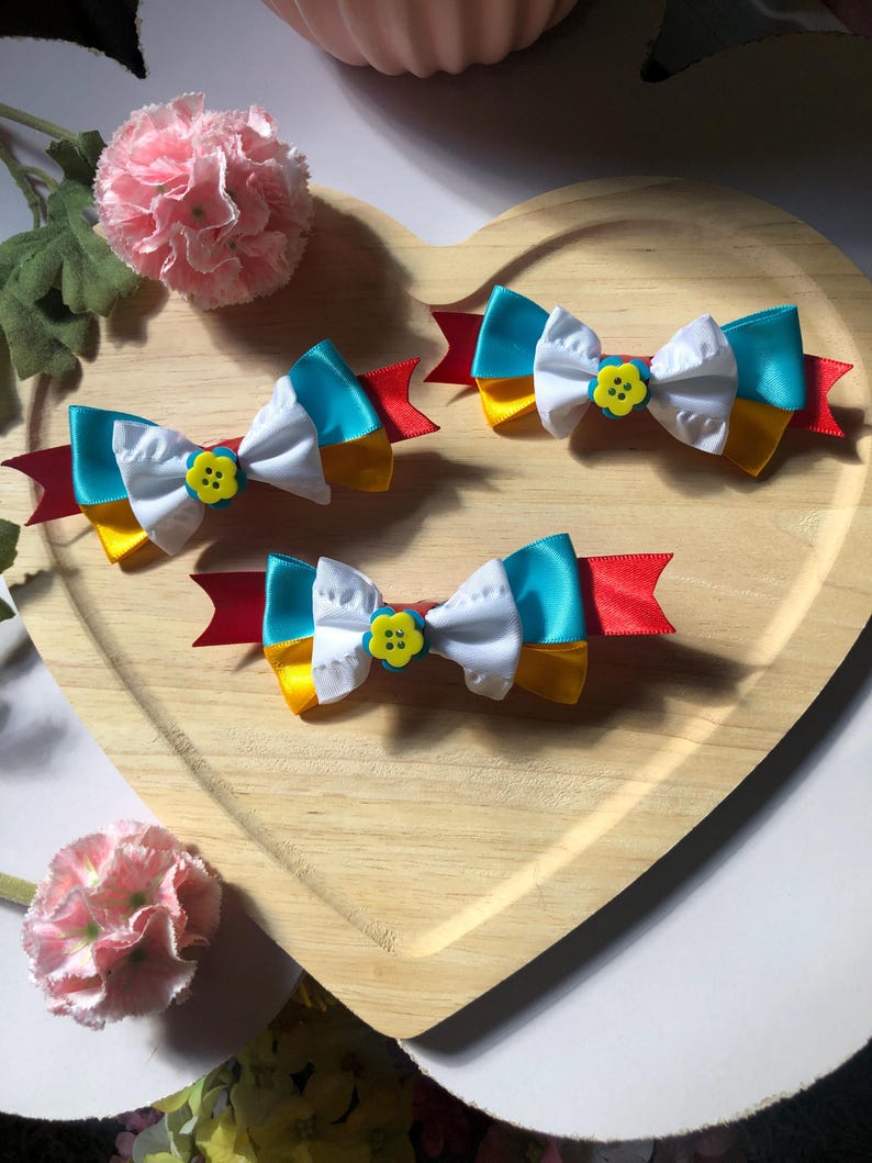 Pu&ograve; includere: Quattro fiocchi per capelli colorati a forma di fiocco, ciascuno con una combinazione di nastri bianchi, gialli e blu turchese, accentuati da una base di nastro rosso e un bottone giallo a forma di fiore. I fiocchi sono disposti su un vassoio di legno a forma di cuore.