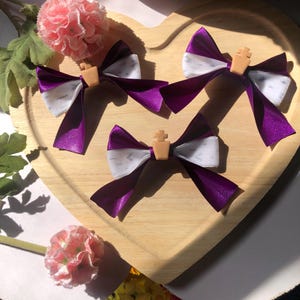 Pu&ograve; includere: Tre fiocchi per capelli in nastro viola e bianco con un piccolo accento a forma di croce color beige sono disposti su un vassoio di legno a forma di cuore. Fiori rosa e verde sullo sfondo.