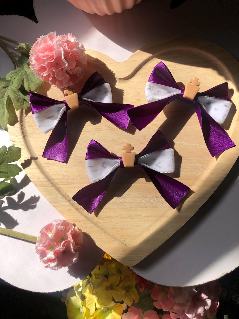 Pu&ograve; includere: Tre fiocchi per capelli in nastro viola e bianco con un piccolo accento a forma di croce color beige sono disposti su un vassoio di legno a forma di cuore. Fiori rosa e verde sullo sfondo.