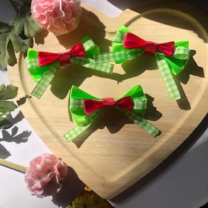 Pu&ograve; includere: Tre fiocchi per capelli con base verde brillante e fiocco centrale rosso sono disposti su un vassoio di legno a forma di cuore. I fiocchi verdi hanno un motivo a quadri bianchi e verdi alle estremit&agrave;. Fiori rosa sullo sfondo.