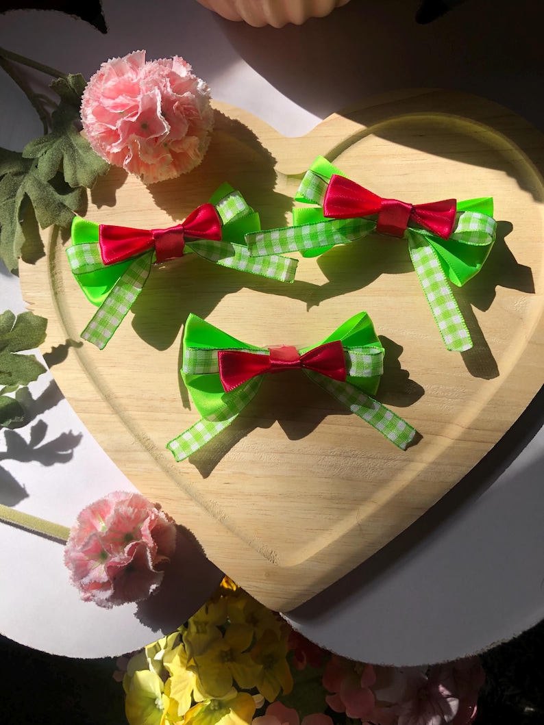 Pu&ograve; includere: Tre fiocchi per capelli con base verde brillante e fiocco centrale rosso sono disposti su un vassoio di legno a forma di cuore. I fiocchi verdi hanno un motivo a quadri bianchi e verdi alle estremit&agrave;. Fiori rosa sullo sfondo.