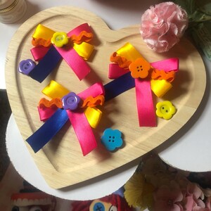 Pu&ograve; includere: Un vassoio di legno a forma di cuore contiene tre fiocchi di nastro colorati con bottoni decorativi. I fiocchi presentano nastri rosa, gialli e blu, accentuati da finiture arancioni e bottoni a forma di fiore. Un garofano rosa e bambole sono sullo sfondo.
