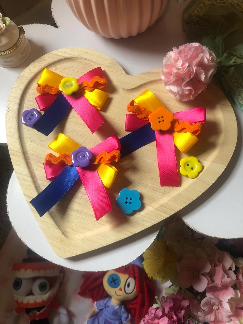 Pu&ograve; includere: Un vassoio di legno a forma di cuore contiene tre fiocchi di nastro colorati con bottoni decorativi. I fiocchi presentano nastri rosa, gialli e blu, accentuati da finiture arancioni e bottoni a forma di fiore. Un garofano rosa e bambole sono sullo sfondo.