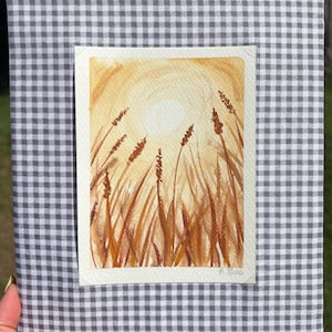 May include: A rectangular notebook or journal cover wrapped in a grey and white gingham fabric. Centered on the front is a watercolor painting depicting a golden sun shining over a field of tall, slender grasses in shades of amber and brown.