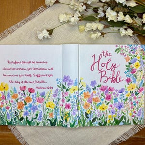 May include: An open book with a floral design. The cover reads "the Holy Bible" in pink script. The interior pages feature a colorful wildflower border and a quote from Matthew 6:34.