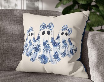 May include: A decorative throw pillow featuring three ghost figures with blue floral patterns and bows. The ghosts have black eyes and are set against a cream-colored background. The pillow has a dark blue border.
