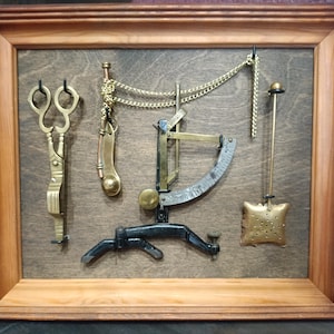 Puede incluir: Un marco que contiene una colección de herramientas antiguas de latón, incluyendo un par de tijeras, un silbato, una balanza, un colador de té y una varilla larga y delgada. Las herramientas están montadas sobre un fondo de madera oscura.