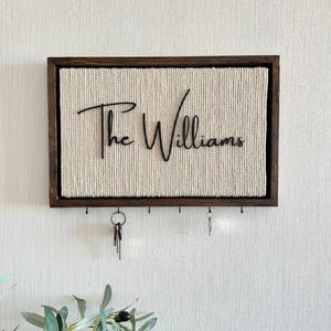 May include: A wooden framed key holder with a woven beige background. The words "The Williams" are written in a black cursive font. Several hooks are below the text, with keys hanging from some of them.