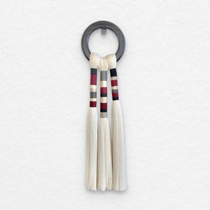 Peut inclure: Suspension murale décorative avec trois glands blancs suspendus à un anneau gris foncé. Chaque gland est orné de bandes rayées rouges, grises et noires. Le fond est un mur blanc uni.